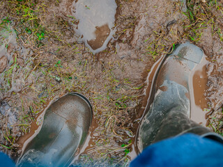 Green hardcore rubber boots in wet brown muddy ground. Top down view. Living in a country rural area in hard conditions concept. - obrazy, fototapety, plakaty