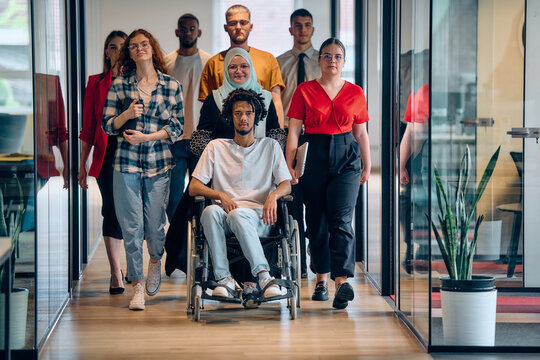 A diverse group of young business people congregates within a modern startup's glass-enclosed office, featuring inclusivity with a person in a wheelchair, an African American young man, and a hijab