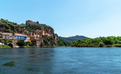 a picturesque riverside village Miravet, Spain. Ancient buildings clinging to the steep hillside, the river flows calmly in the foreground, framed by the lush greenery of the surrounding landscape.