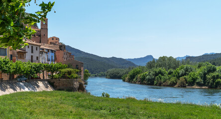 Nestled by a serene river, ancient terracotta-roofed houses bask under a clear sky, framed by...