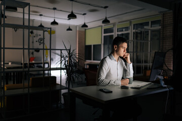 Depressed remote view of focused millennial male hard working from office, thinking and feeling thoughtful, overload at night, sitting on desk at late night in dark room. Concept overwork and deadline