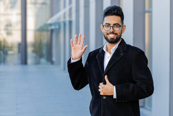 Hello. Indian businessman smiling friendly at camera, waving hands gesturing hi, greeting or...