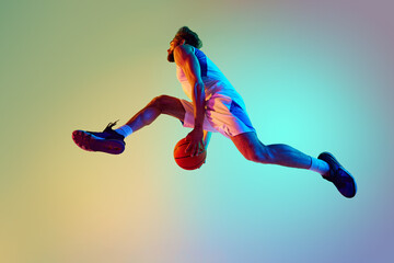Slam dunk. Bottom view of bearded young man, basketball player in motion, jumping with ball against gradient background in neon light. Concept of sport, competition, active and healthy lifestyle, game