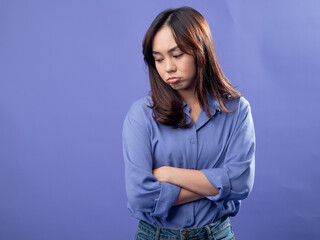 A young Asian woman in a light blue button-up shirt and jeans stands against a solid lavender background with her arms crossed and a sad, pouty expression, indicating disappointment or sorrow.