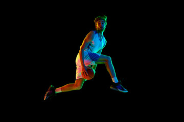 Fototapeta na wymiar Dynamic image of young man, basketball player in mid-air, training, jumping with ball against black background in neon light. Concept of sport, competition, active and healthy lifestyle, game