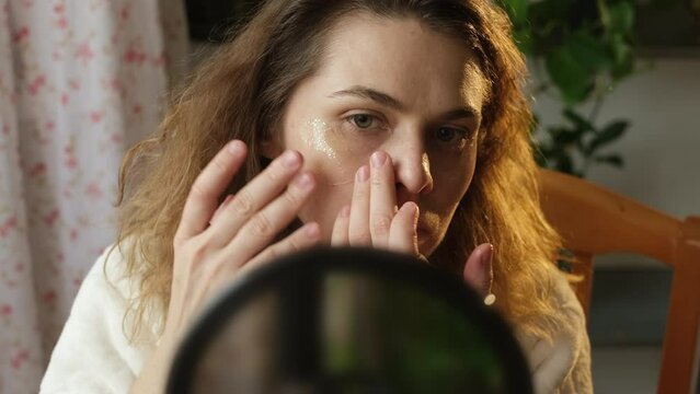 Woman without make up applying cosmetic patches under eyes looking in mirror.