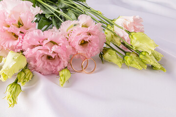 Two gold classic wedding rings on a white satin background with fresh pink austoma flowers. A copy...
