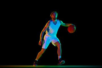 Young athletic man, basketball player in motion with ball, training against black background in neon light. Concept of sport, competition, active and healthy lifestyle, game