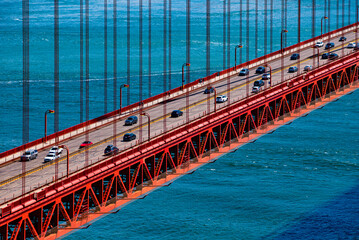 Bridge deck of iconic Golden Gate Bridge with famous 4 lane coastal “Highway 1“ and cars. Part of red steel construction with cars and blue wavy San Francisco strait in the background on a sunny day.