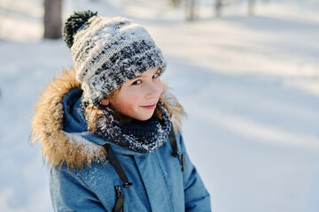 Cute smiling boy in grey knitted beanie hat and blue winter jacket looking at camera while walking along road in park or forest