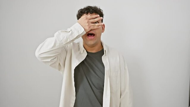 Shocked young arab man in casual clothes, peeking through fingers covering face, afraid, on isolated white background
