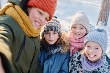 Obraz premium One of four happy kids taking selfie on smartphone while standing next to his cheerful friends wearing warm winterwear