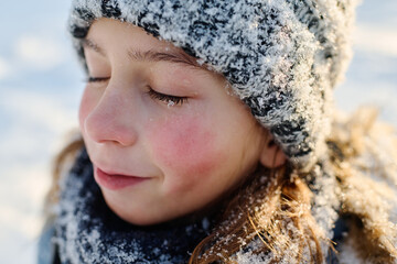Close-up portrait of cute boy with closed eyes and snowflake on eyelashes standing in front of camera on winter day and enjoying stroll