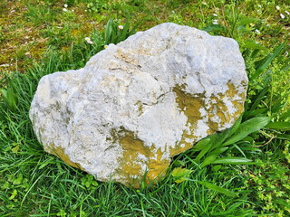 Großer, unregelmäßig geformter Stein, der auf einem Grasfeld liegt. Der Stein hat eine raue,...