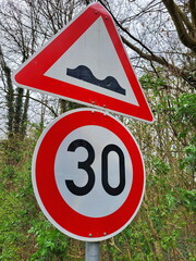 Verkehrsschilder an einer Straße. Das obere Schild ist ein Warnschild, das auf eine unebene...