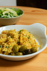 Bacalhau à Brás, a traditional Portuguese dish, served on a white plate in a restaurant setting. The shredded cod is mixed with eggs, crispy potatoes, parsley, and black olives.
