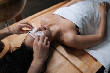 Closeup top view of spa master applying cotton pad to serene female client eyes in preparation for...