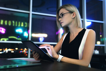 Fully concentrated beautiful young businesswoman with blonde hair glasses spectacles holding tablet in hand sitting on a table at coworking with look in laptop. Female entrepreneur using gadget - Powered by Adobe
