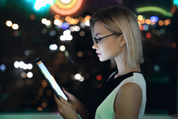 Side view horizontal shot of attractive female businesswoman ceo entrepreneur student in glasses that reading watching checking message news task late night in office building standing against window