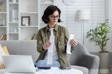 Worried and serious young woman sitting on sofa at home, checking receipts for purchases and...