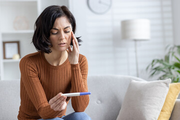 Close-up photo of an upset and worried woman sitting on the sofa at home and looking disappointedly at the one-time fake pregnancy test she is holding in her hands - obrazy, fototapety, plakaty