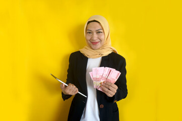 Adult Asian muslim businesswoman holding tablet and money with happy expression