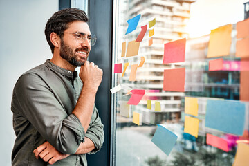 Planning goals at work. Portrait of confident smiling adult man wearing glasses standing near a glass wall with colorful stickers and looking at them.Sticky note on glass wall. Copy space.