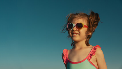 Portrait of laughing girl of 6 years in blue swimsuit and heart-shaped sunglasses against blue sky,...
