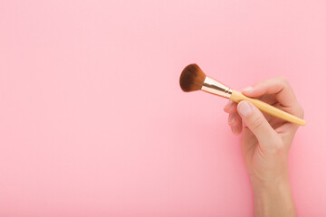 Young adult woman hand fingers holding and showing new big makeup brush with soft bristles on pink table background. Pastel color. Female beauty product. Closeup. Empty place for text. Top down view. - 794005779