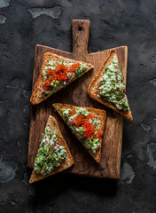 Delicious tapas, appetizer, snack - bruschetta with avocado, eggs, gherkins, greens spread and red caviar on a wooden chopping board on a dark background - 794006965