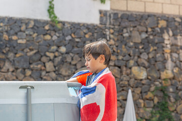 Young boy drying off with towel by the pool, pensive expression, outdoor summer day.