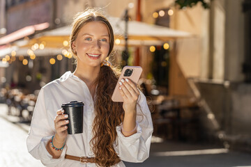 Happy redhead teenager girl enjoying morning coffee hot drink and smiling outdoors. Relaxing taking a break. Woman walking in urban city sunshine street, drinking coffee to go. Town lifestyles outside