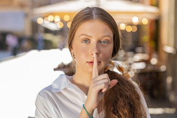 Shh be quiet please. Teenager redhead girl presses index finger to lips makes silence hush gesture sign do not tells gossip secret outdoors. Young woman walking in urban city street. Town lifestyles
