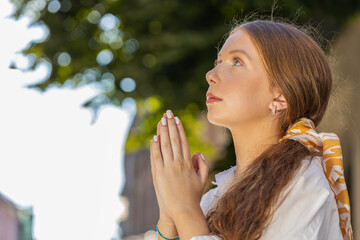 Portrait of redhead religion teenager girl praying closed eyes to God asking for blessing, help, forgiveness outdoor. Young woman clasping hands wishing luck in urban sunny city street. Town lifestyle