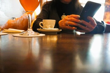 Frau sitzt mit Handy in Händen vor braunem Holztisch mit Cocktailglas und weißer Tasse Kaffee in...