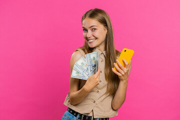 Young woman looking smartphone display sincerely rejoicing win, receiving money dollar cash banknotes, celebrating success lottery luck. Girl isolated alone on pink studio background. Lifestyles