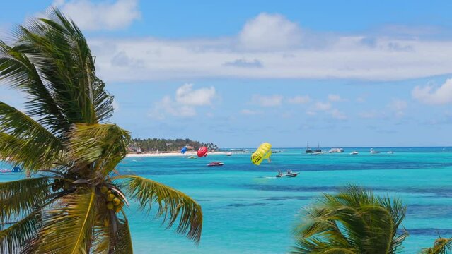 View through palm trees to a large white sand beach with many resorts and hotels on the coast of turquoise Caribbean sea. Watersports excursions while summer vacations in Punta Cana