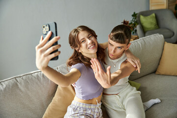 Two women in comfy attires smiling while taking a selfie on a couch.
