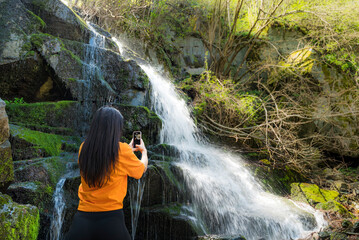 Woman standing in front of waterfall and taking pictures with the phone 