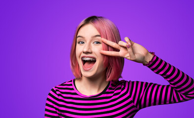 Photo image - happy pink beautiful woman in braces show v-sign victory peace hand gesture two fingers, with opened mouth, isolated violet purple background. Girl at studio. Dental care ad concept