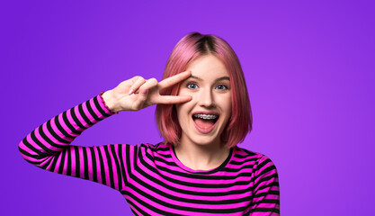 Photo image - happy pink beautiful woman in braces brackets show v-sign victory peace hand gesture two fingers, opened mouth, isolated violet purple background. Girl at studio. Dental care ad concept