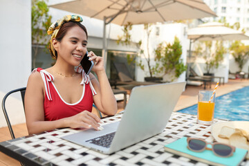 Smiling businesswoman in bathing suit sitting at table next to swimming pool, talking on phone and working on laptop - Powered by Adobe