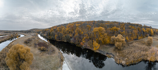 Aerial wide panorama of river valley with golden autumn forest and grey cloudy sky in Ukraine