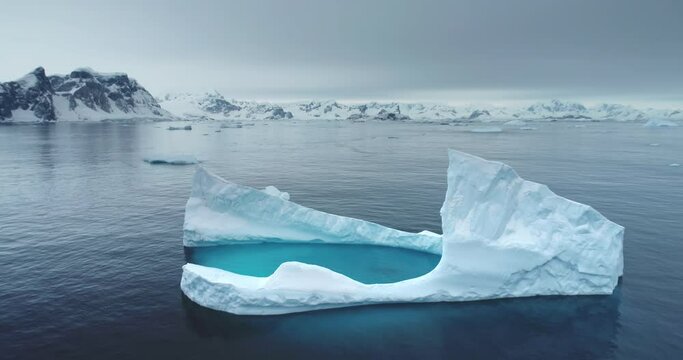 Antarctic melting iceberg floating in cold blue water, aerial. Snow-covered mountains in background. Melting glacier drift in Polar ocean. Ecology, melting ice, climate change, global warming concept