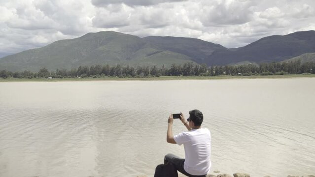 hiker sitting in front of a lake and a mountain in a valley, taking photos with his cell phone