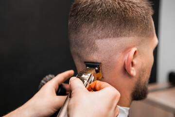 Master cuts customer hair using comb and trimmer in barbershop closeup. Male client sits in front of mirror while barber cuts hair in salon