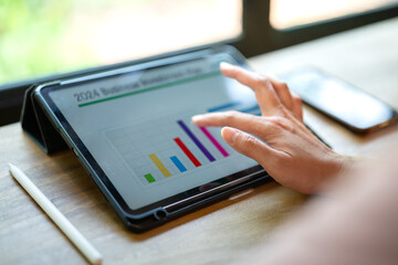 Action of a person hand is touching on tablet screen to review the 2024 business investment plan with graph and chart data. Business working with technology concept, close-up and selective focus.	
