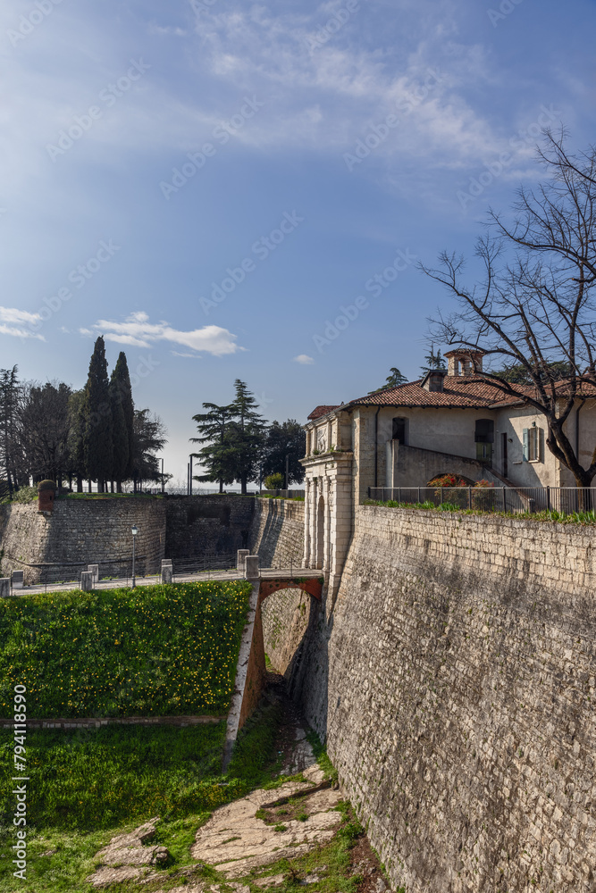 Wall mural This vertical shot captures the charm of Brescia castle entrance with its arched bridge, set against a backdrop of verdant trees and a sprawling blue sky