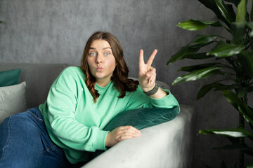Portrait of young woman looking at camera and relaxing on a sofa after work at home. People lounge concept. 30s girl showing peace sign with fingers