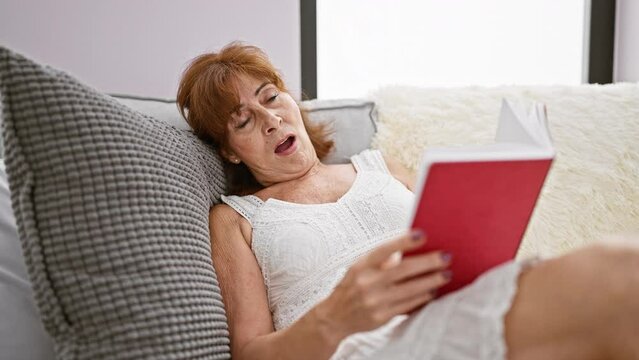 Mature redhead woman yawning while reading a red book, about to nap on a cozy sofa at home, depicting everyday relaxation and senior lifestyle.